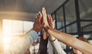Five hands high-fiving in an open-plan office with sun coming in the back windows.
