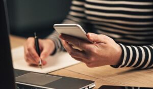 A virtual assistant holding mobile and writing down something with a laptop in front of her while using her favourite apps.