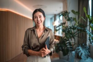 Positioned within a modern corporate environment, a mid-aged Asian businesswoman of striking beauty stands in her office, poised with a digital tablet, using it to execute digital tools and wireless technology to perfection in her strategic endeavors.